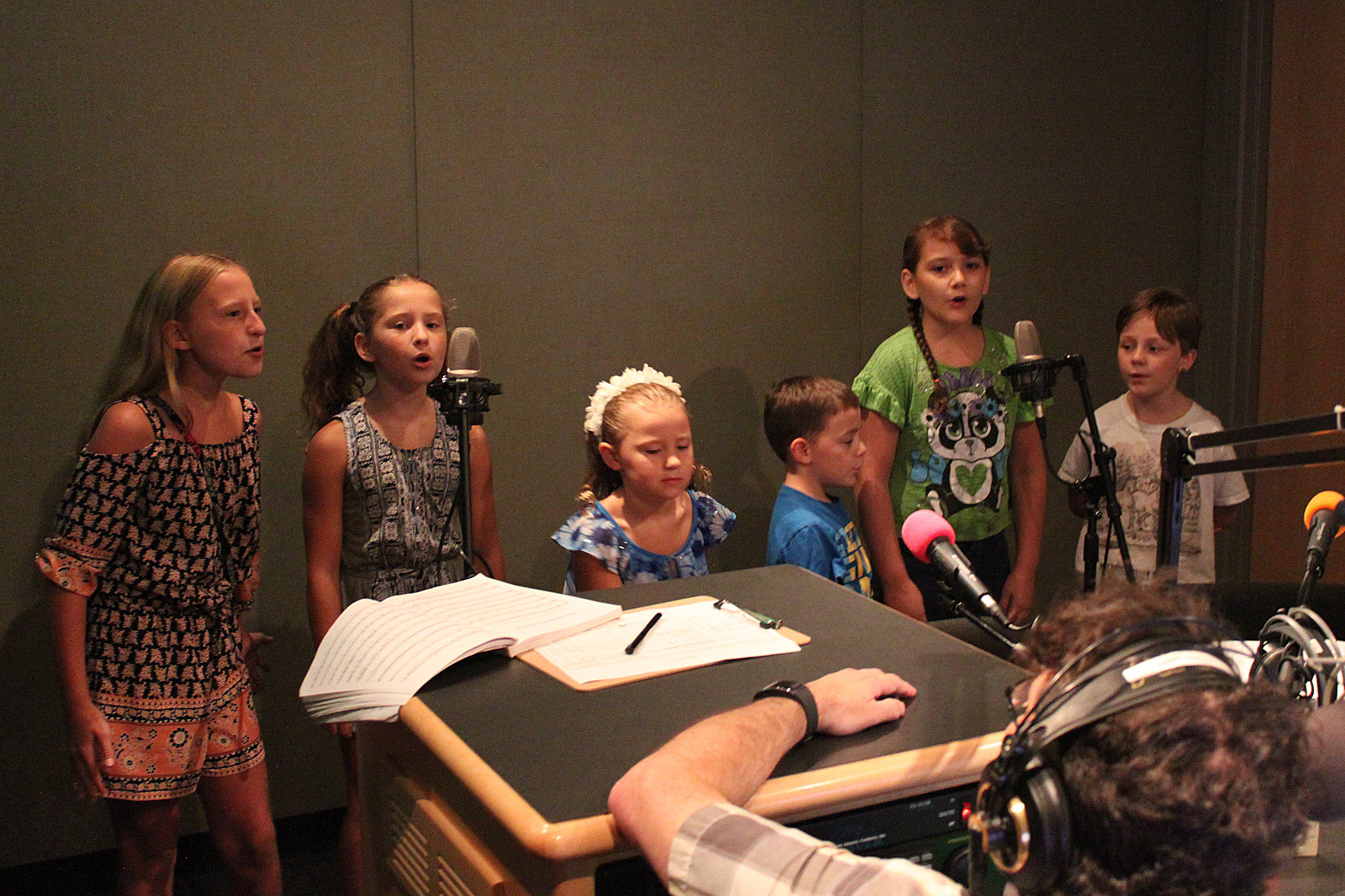 Six youth singing into microphones as an adult wearing headphones sitting behind a microphone listens in a radio studio