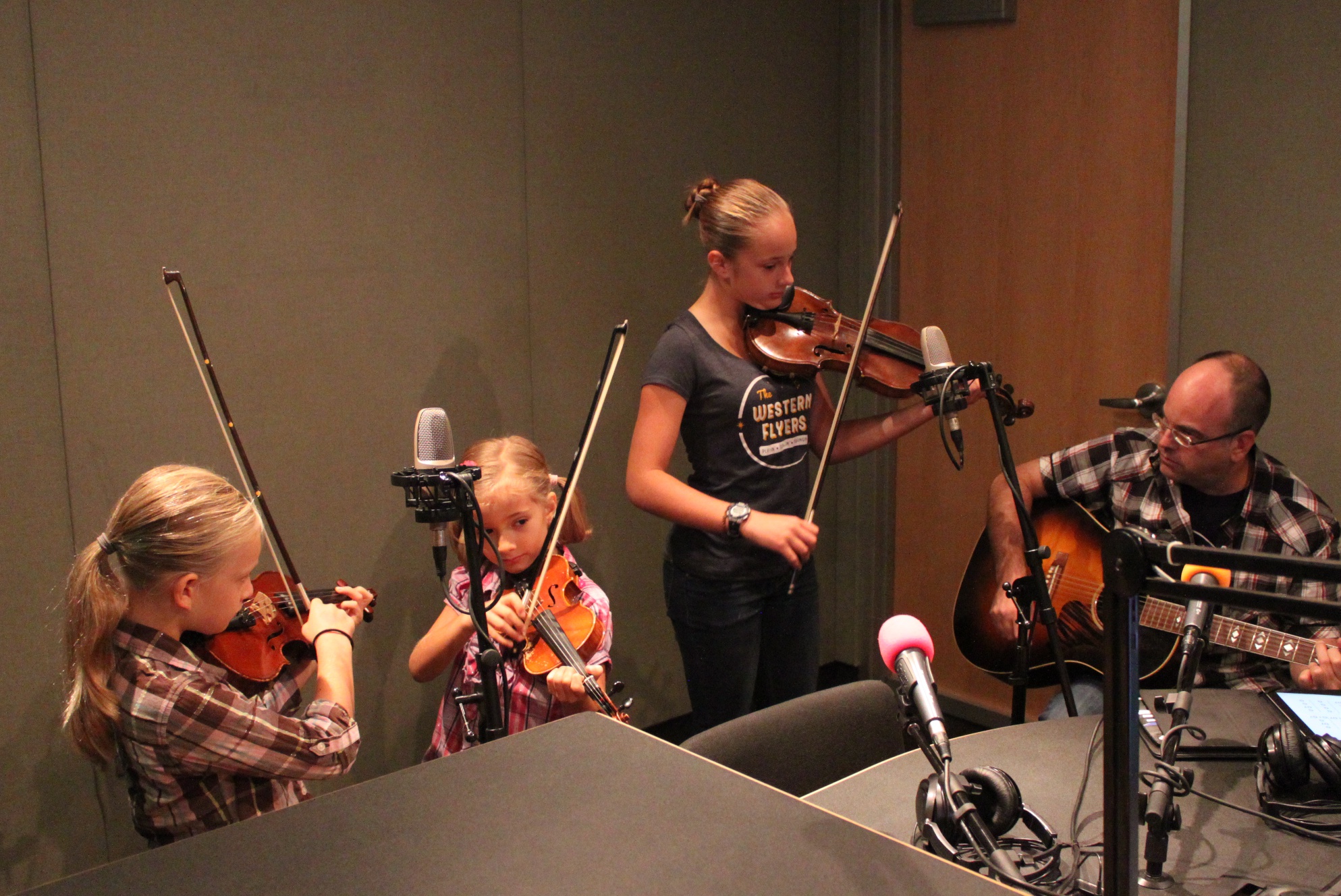 Two youth playing violin, an teen playing violin, and an adult playing acoustic guitar in a radio studio