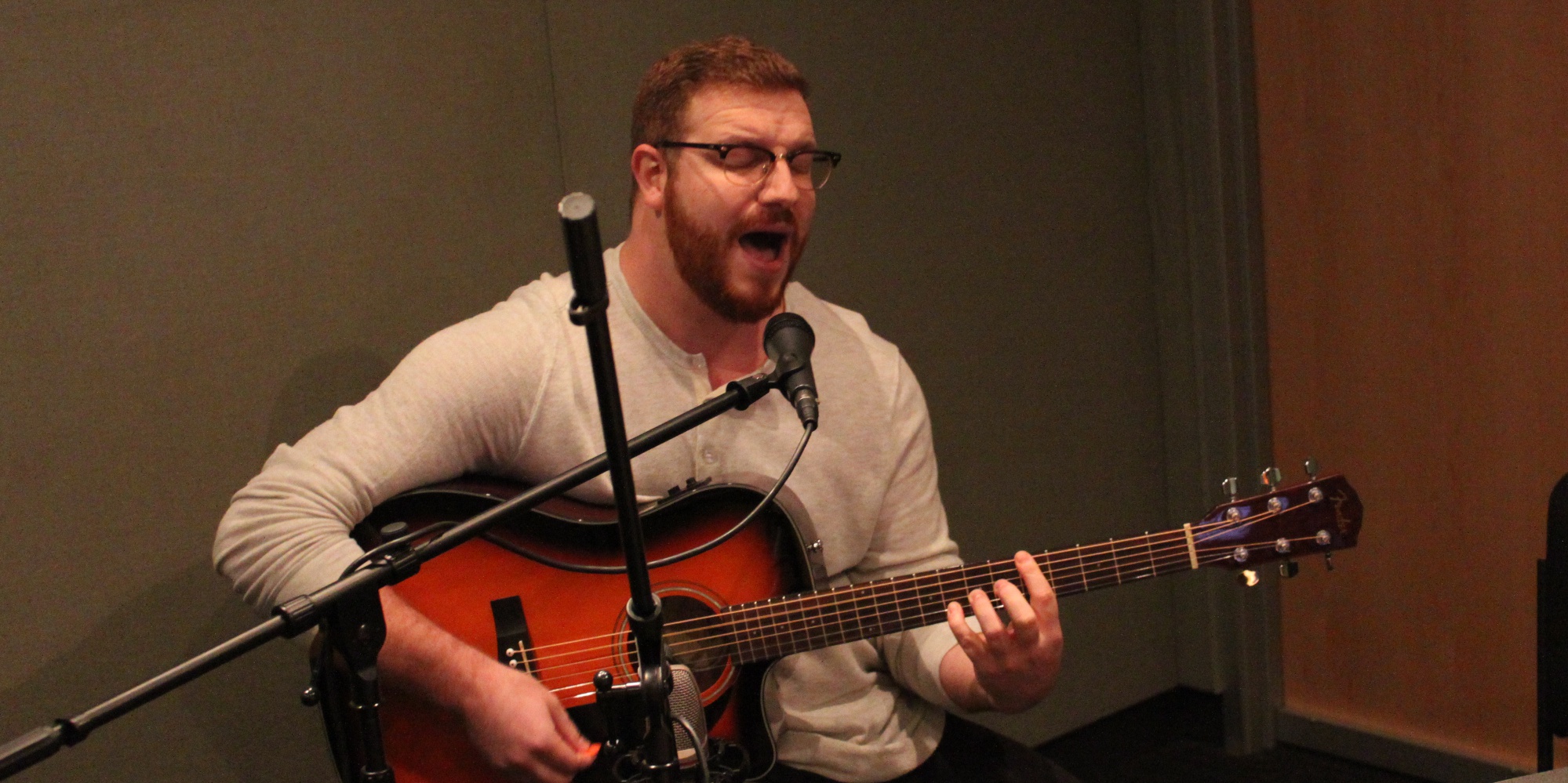 An adult playing acoustic guitar and singing into a microphone in a radio studio