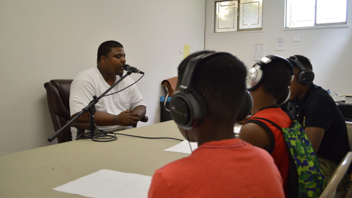 An adult speaking into a microphone being interviewed by three youth wearing headphones