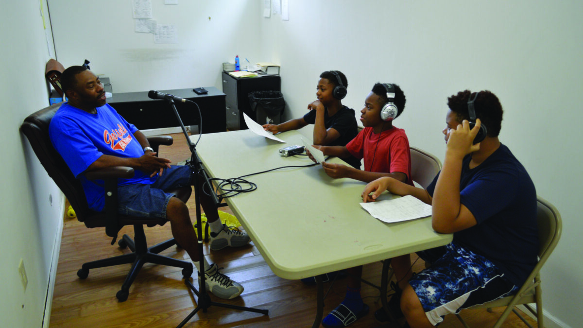 An adult speaking into a microphone connected to a portable recorder being interviewed by three youth wearing headphones