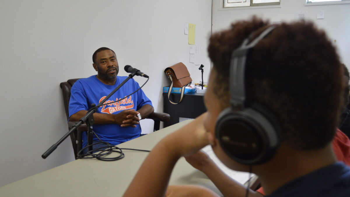 An adult speaking into a microphone being interviewed by a youth wearing headphones