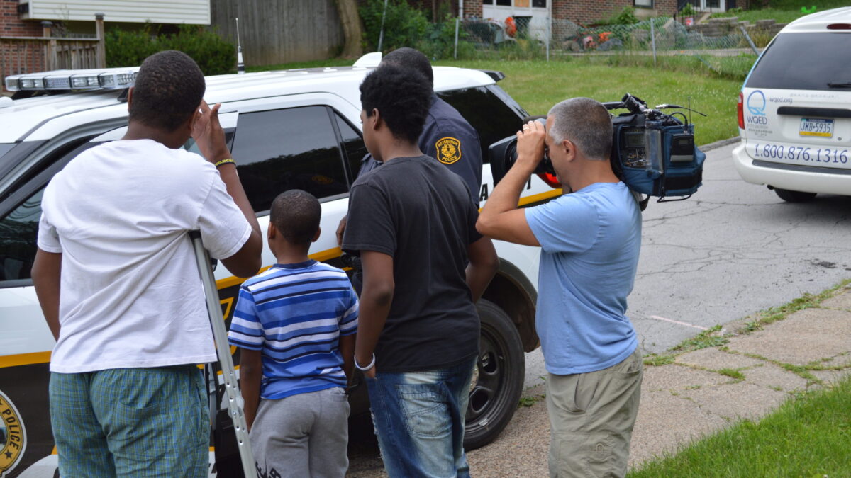 Three youth, an adult holding a camera, and a police officer, all looking inside a police officer