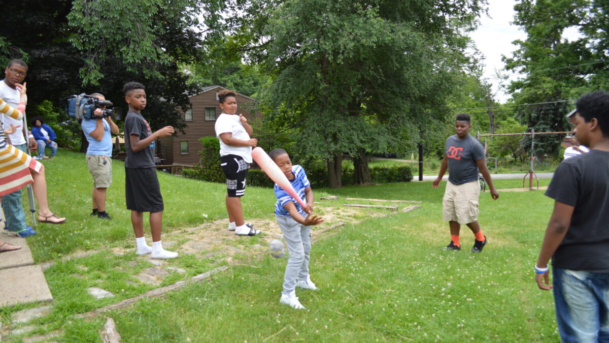 Five youth playing with a baseball bat in a green field as an adult holding a large video camera films