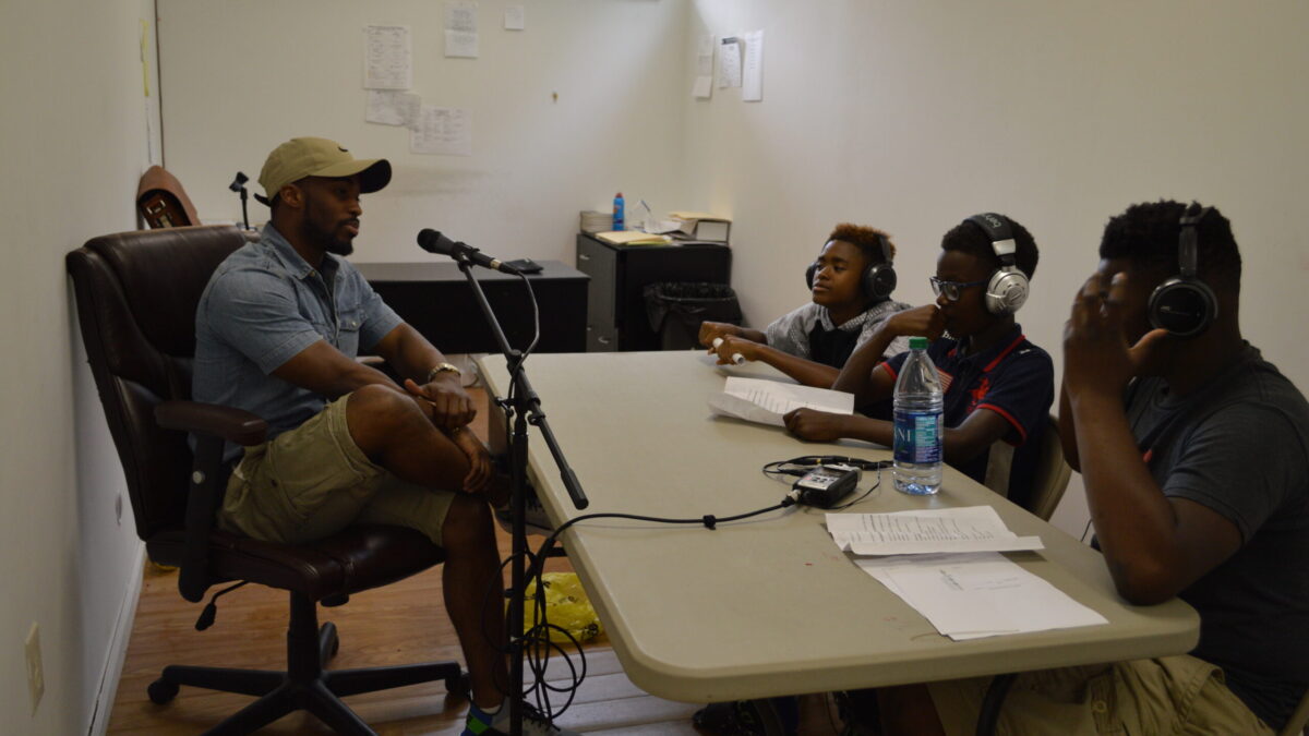 An adult speaking into a microphone connected to a portable recorder being interviewed by three youth wearing headphones