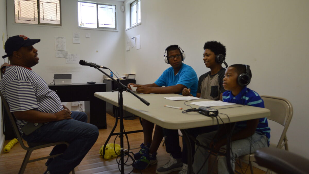 An adult speaking into a microphone connected to a portable recorder being interviewed by three youth wearing headphones
