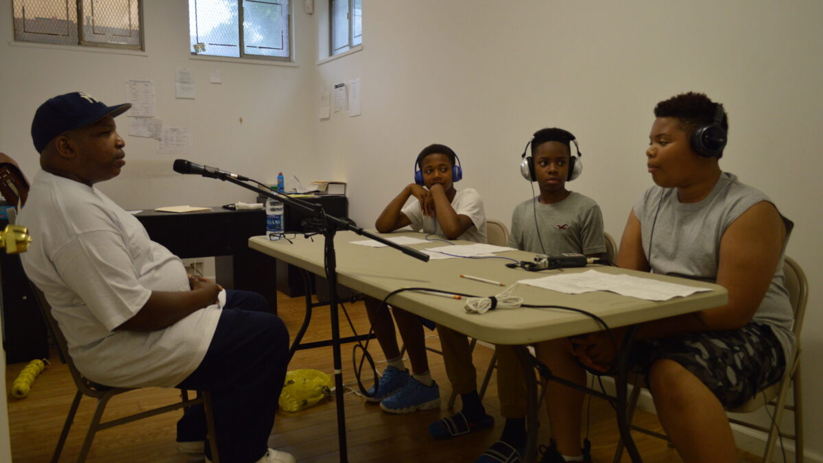 An adult speaking into a microphone connected to a portable recorder being interviewed by three youth wearing headphones
