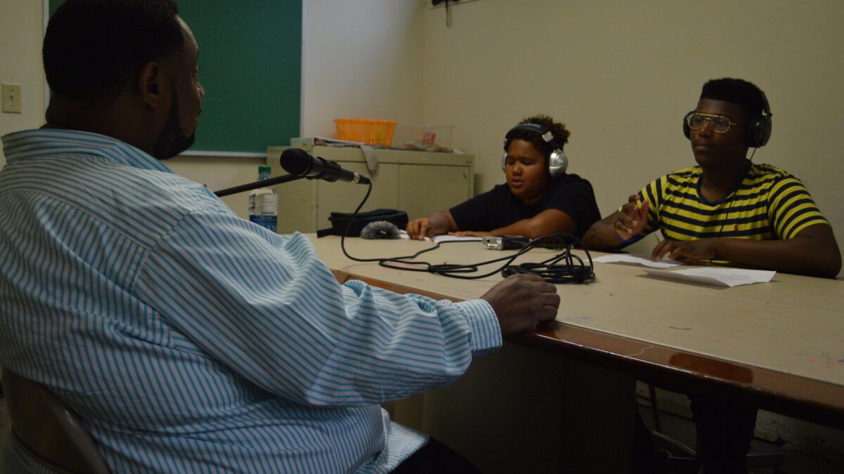 An adult speaking into a microphone connected to a portable recorder being interviewed by two youth wearing headphones