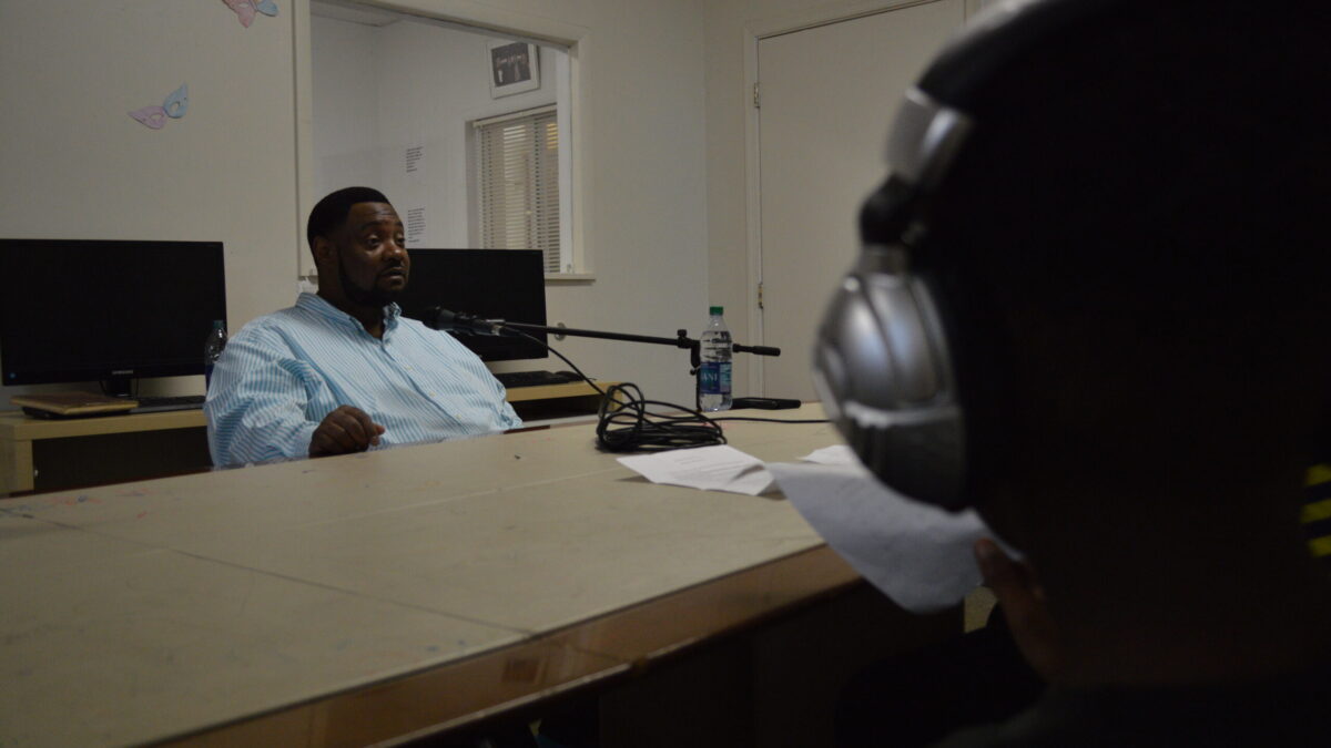 An adult speaking into a microphone connected to a portable recorder being interviewed by a youth wearing headphones
