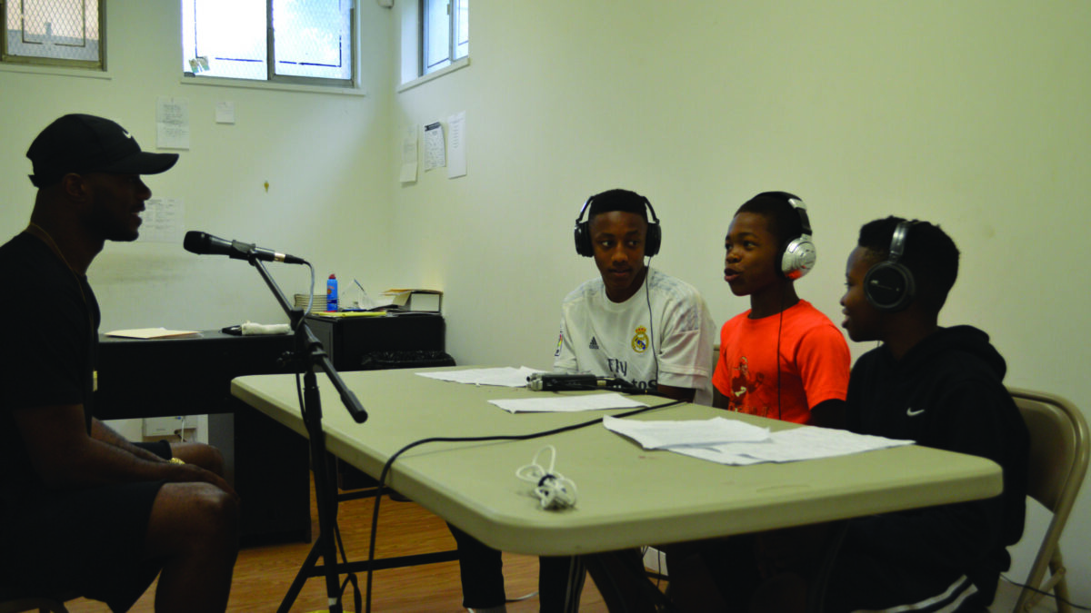 An adult speaking into a microphone connected to a portable recorder being interviewed by three youth wearing headphones