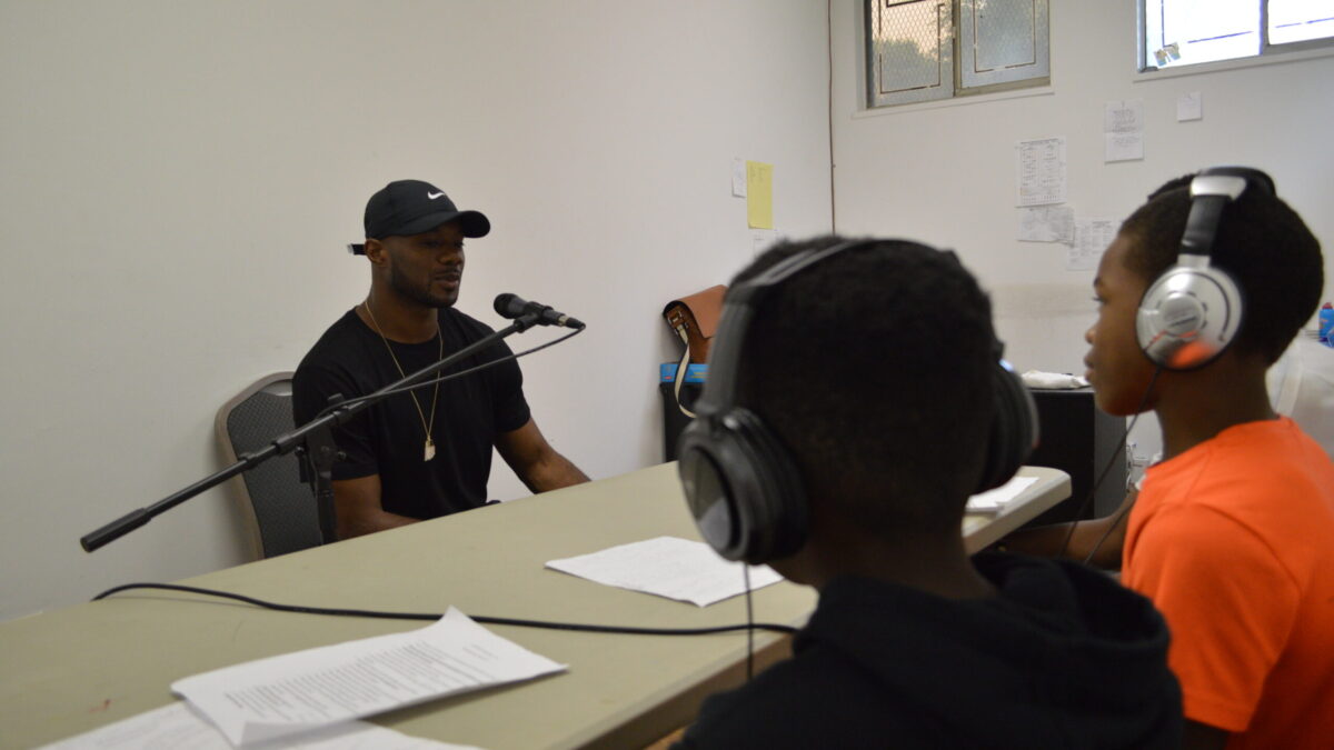 An adult speaking into a microphone connected to a portable recorder being interviewed by two youth wearing headphones