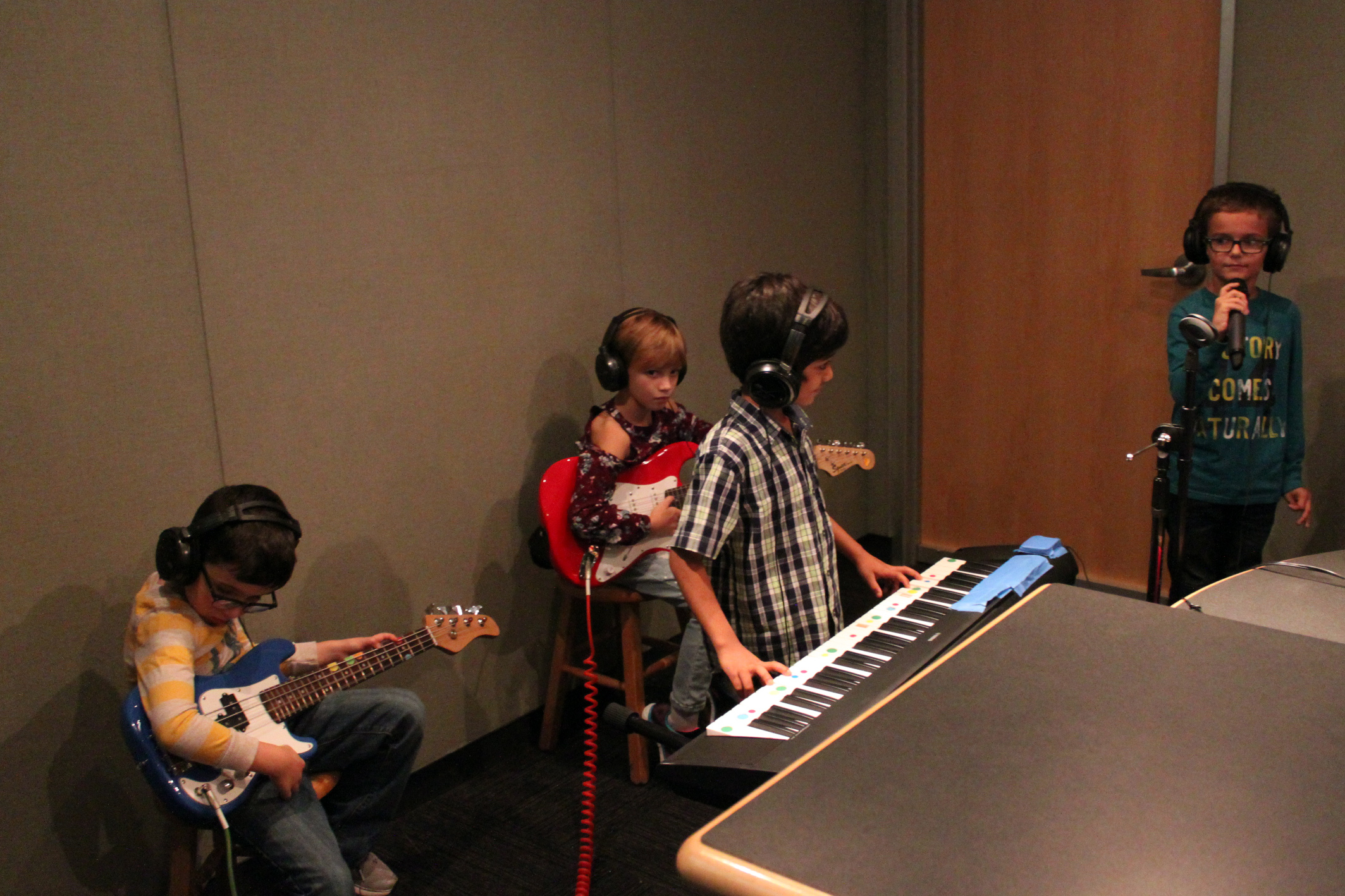 Four youth wearing headphones in a radio studio, one playing electric guitar, one playing electric bass, one playing a piano keyboard, and one singing into a microphone