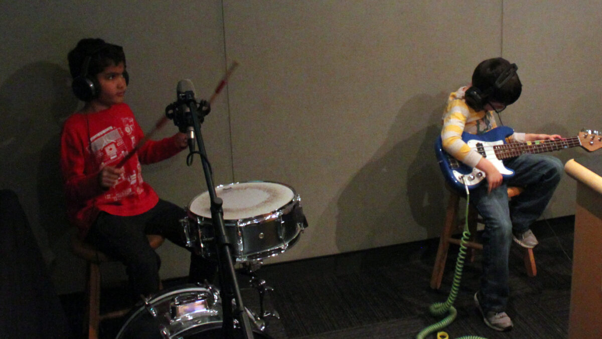 Two youth wearing headphones in a radio studio, one playing drum set, the other playing electric bass