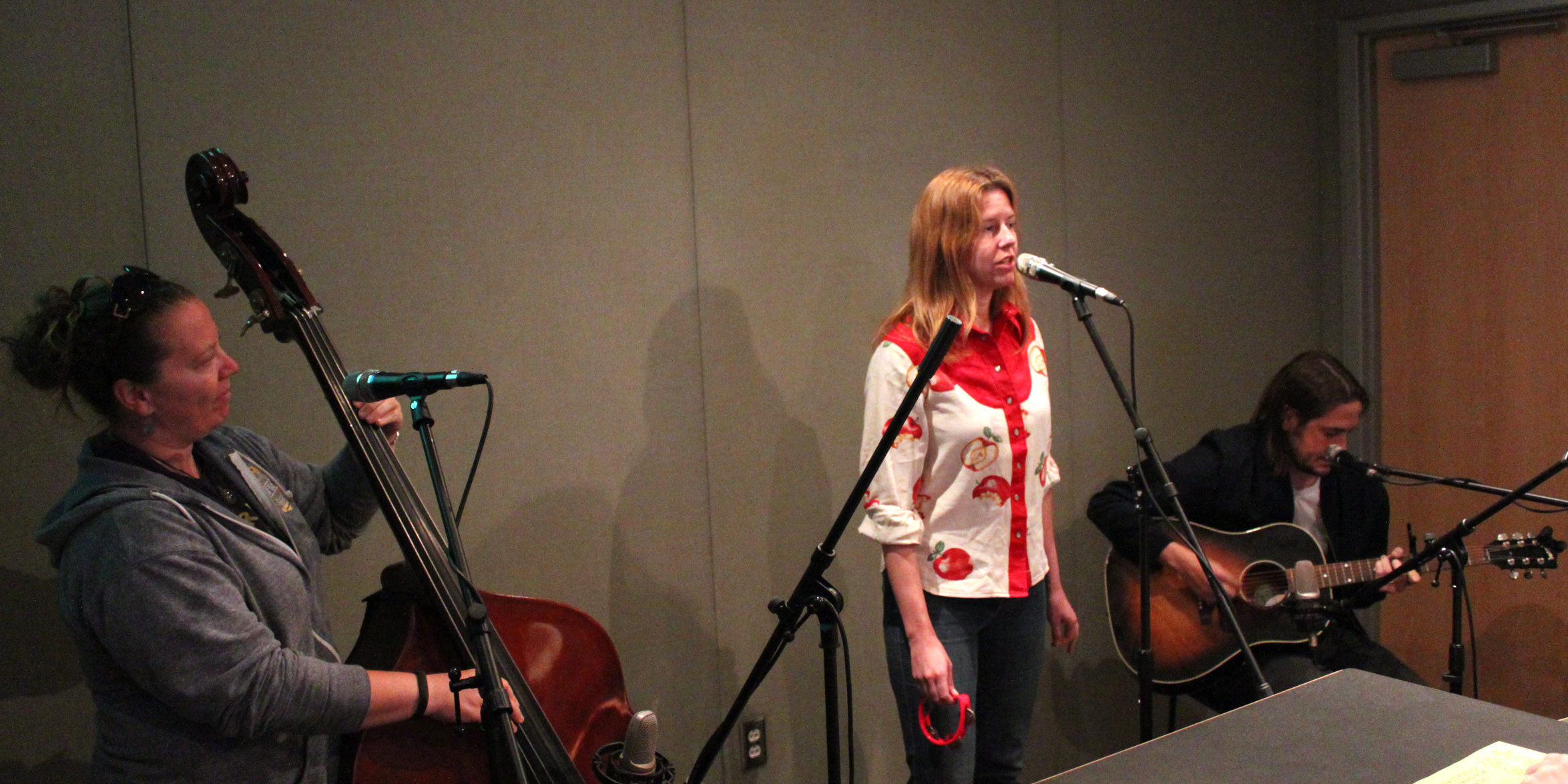 An adult playing upright bass, an adult singing into a microphone, and an adult playing acoustic guitar in a radio studio