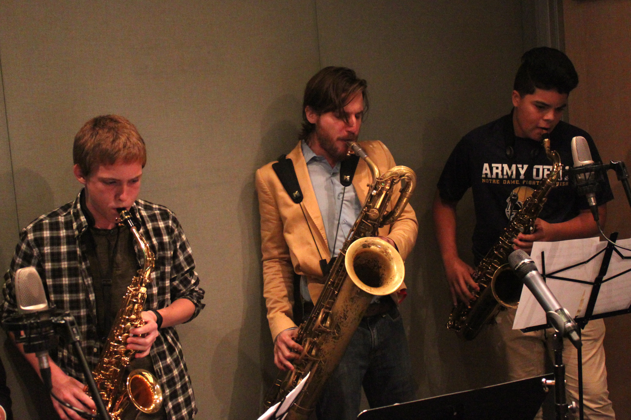 An adult playing a baritone saxophone, a teen playing tenor saxophone, and a teen playing an alto saxophone, all looking at sheet music on music stands in a radio studio