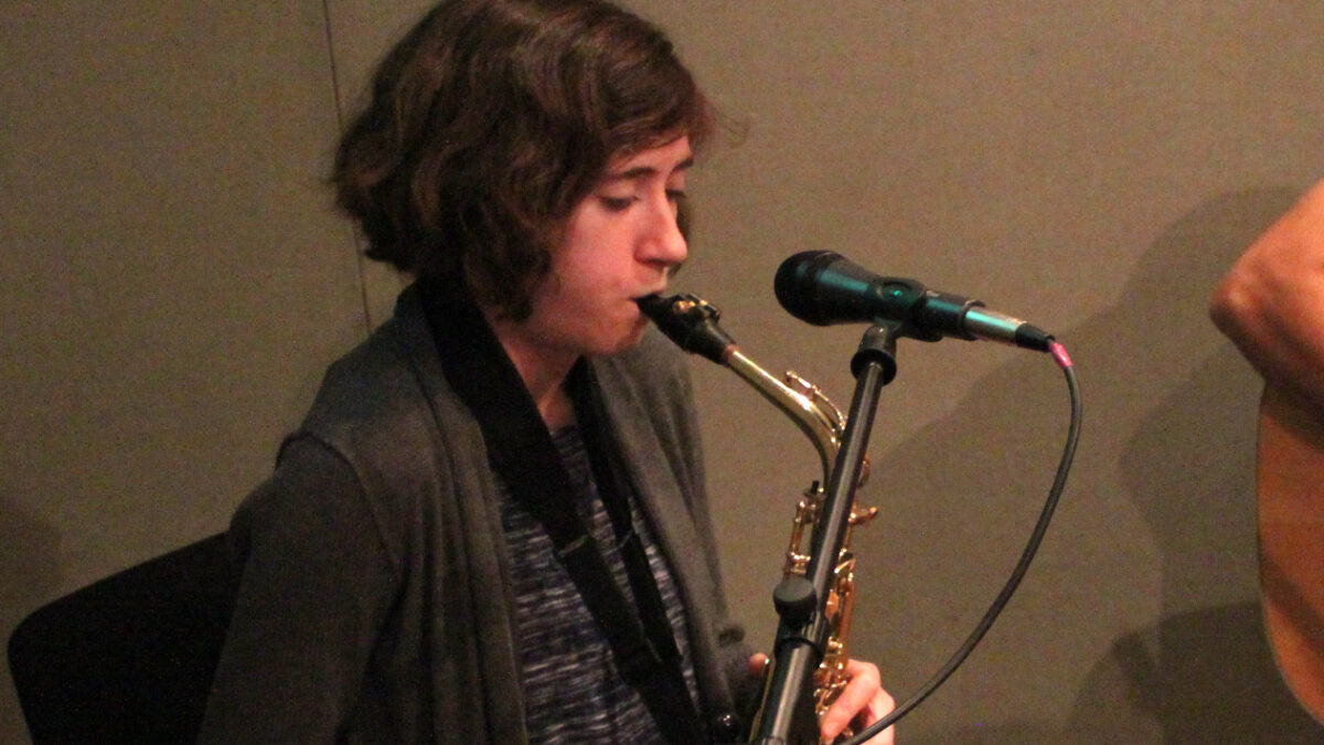 An adult playing alto saxophone sitting behind a microphone in a radio studio