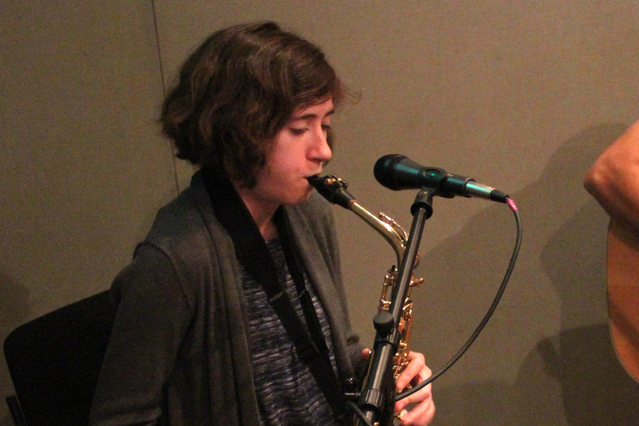 An adult playing alto saxophone sitting behind a microphone in a radio studio