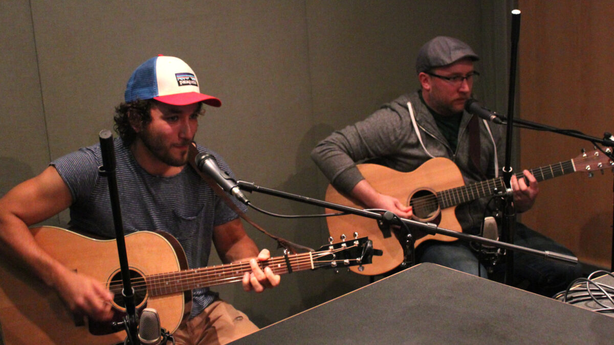 Two adults playing acoustic guitars and singing into microphones in a radio studio