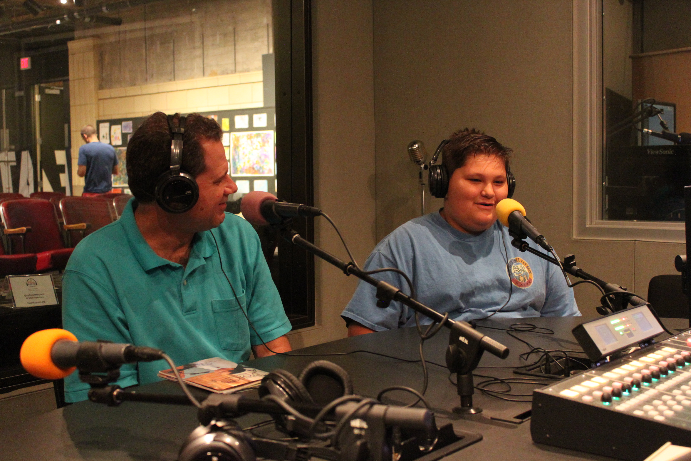 An adult and a youth wearing headphones speaking into microphones in a radio studio
