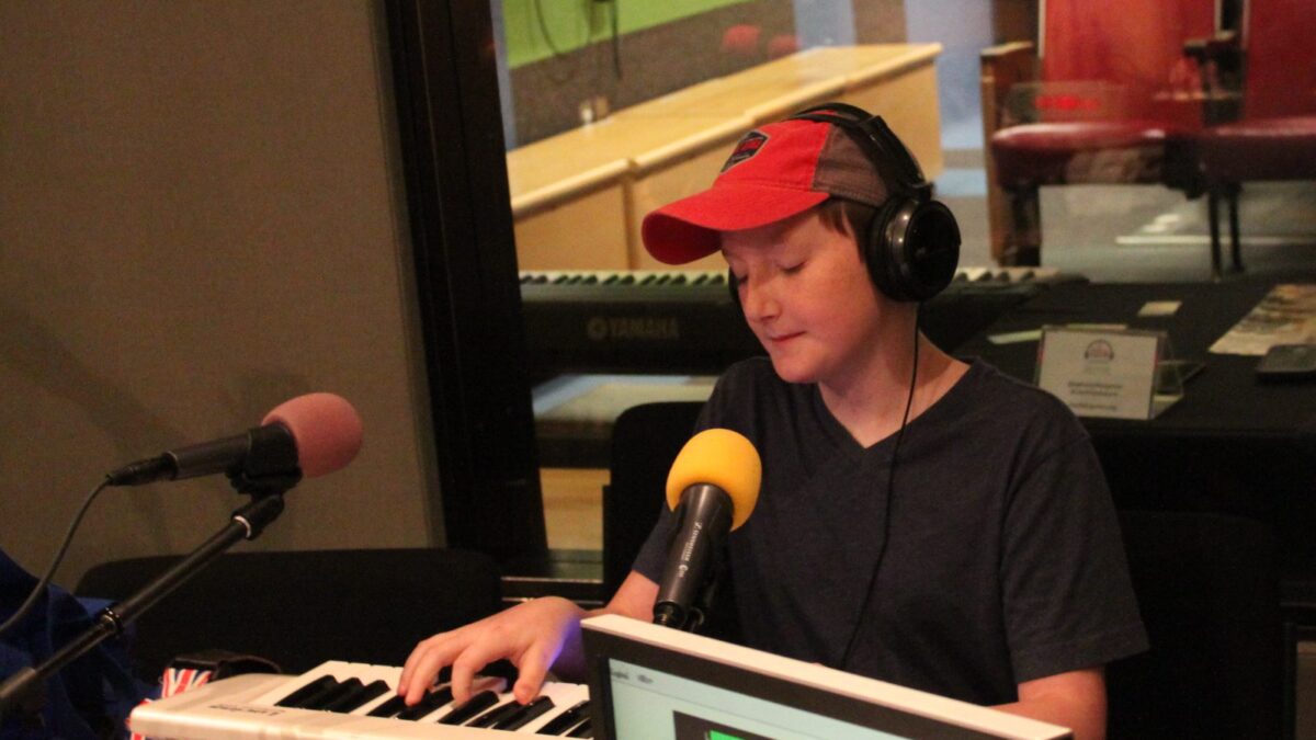 A teen wearing headphones playing a piano keyboard sitting behind microphones in a radio studio.