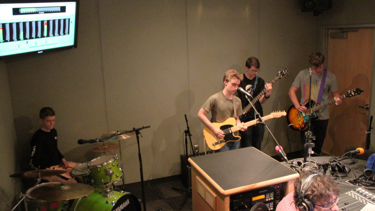 A teen playing drum set, a teen playing electric bass, and two teens playing electric guitar, one singing into a microphone, as an adult wearing headphones listens in a radio studio