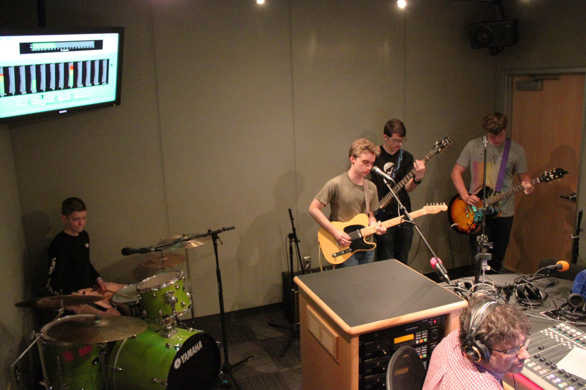 A teen playing drum set, a teen playing electric bass, and two teens playing electric guitar, one singing into a microphone, as an adult wearing headphones listens in a radio studio