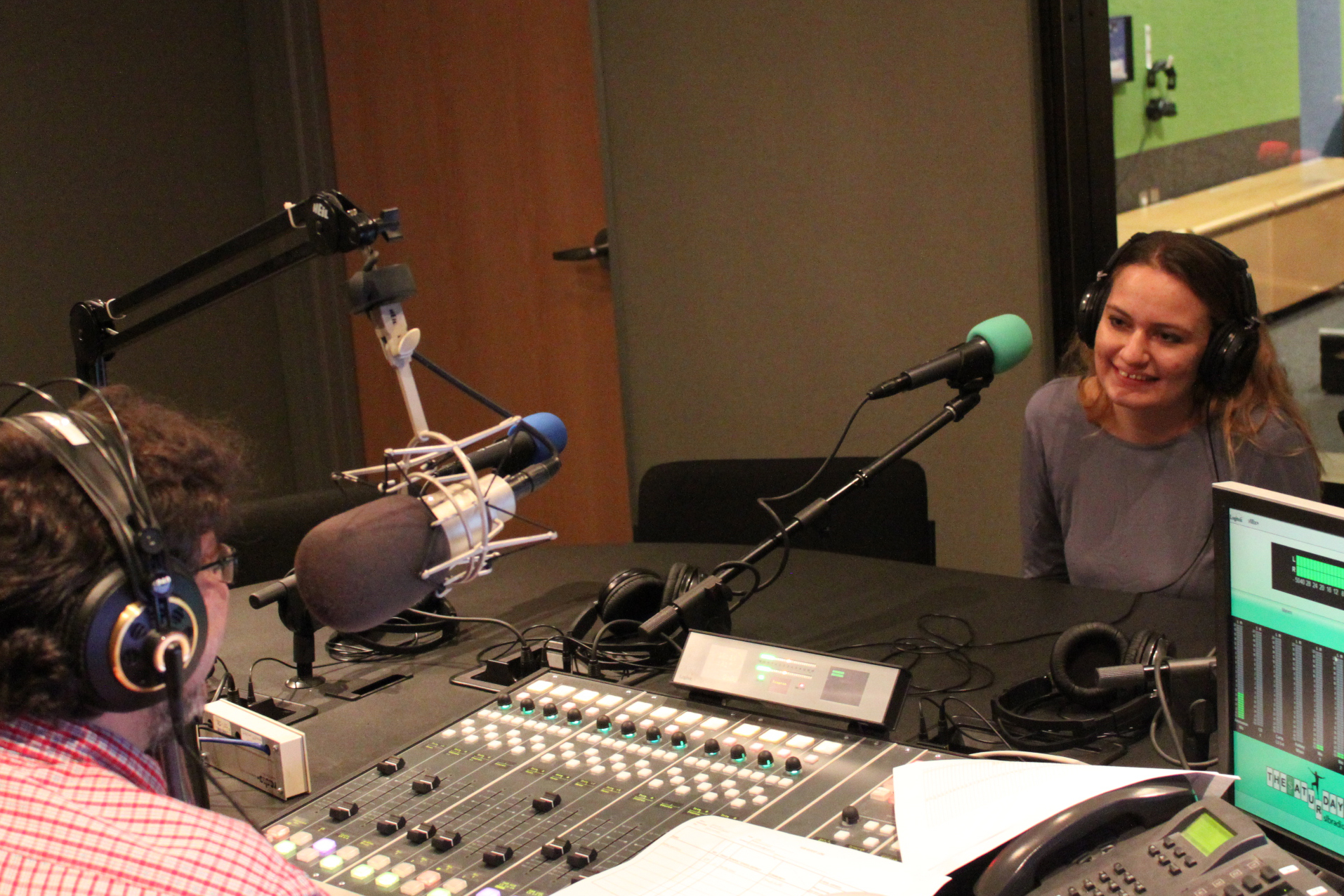 An adult sitting behind a microphone being interviewed by an adult wearing headphones sitting behind a microphone and a mixing board in a radio studio