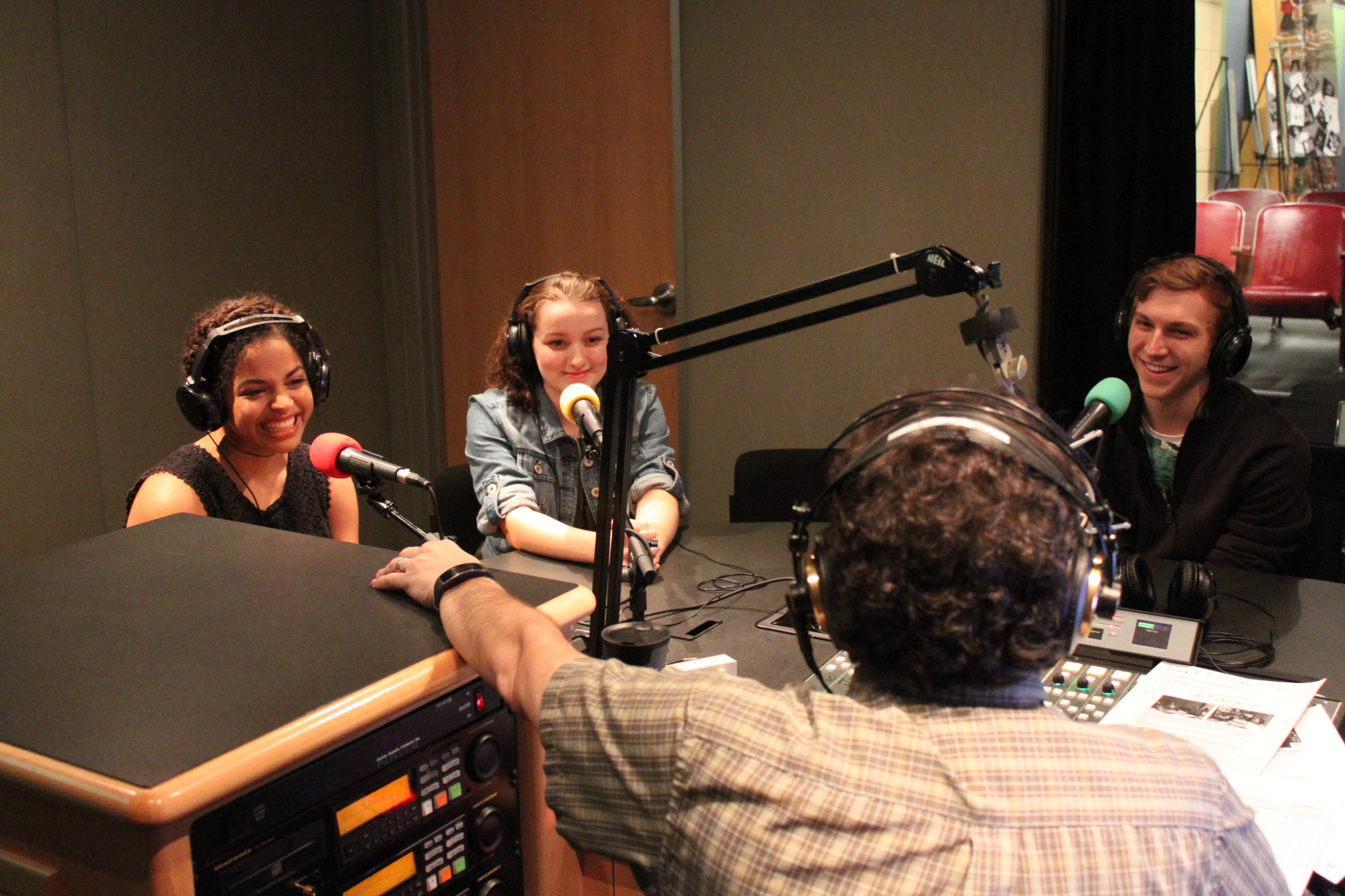 Three teens wearing headphones sitting behind microphones being interviewed by an adult wearing headphones sitting behind a microphone and a mixing board in a radio studio