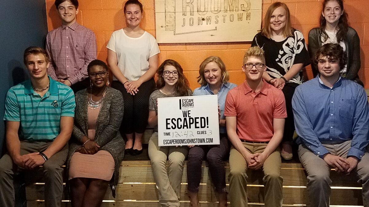 Ten teens sitting on a set of stairs, two holding a sign declaring that they escaped