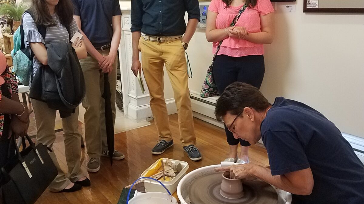 Four teens looking at an adult at a pottery wheel molding a piece of clay