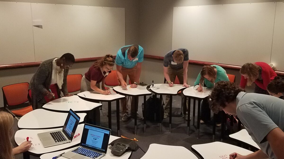 Eight individuals writing on pieces of paper around a collection of desks arranged in a circle