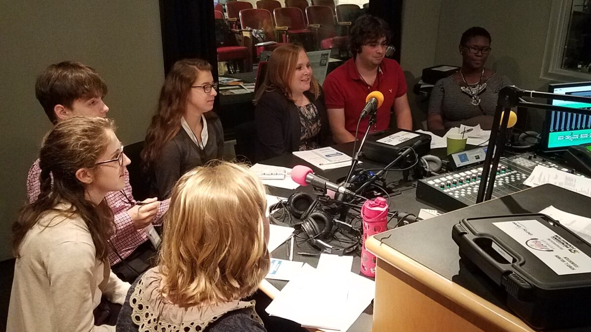 Seven teens sitting around microphones and pieces of paper in a radio studio