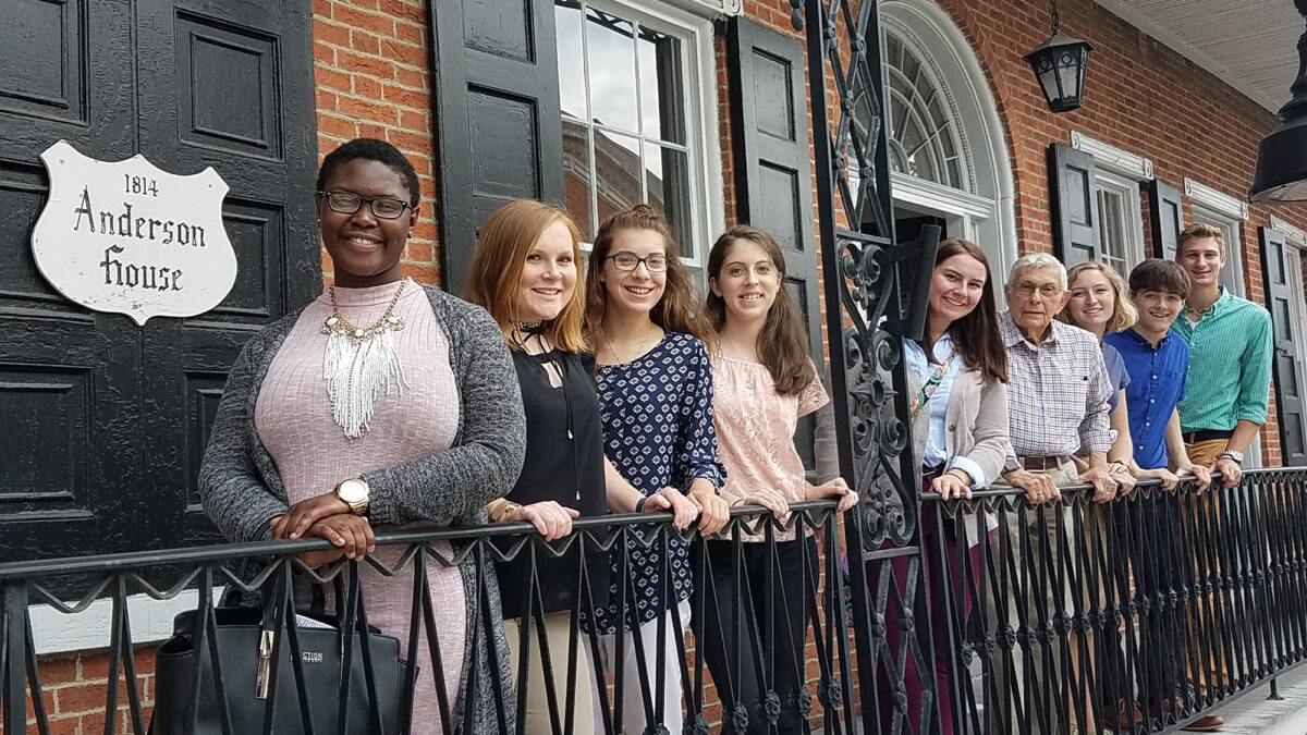 Eight teens and one adult posing in front of an old fashioned brick house