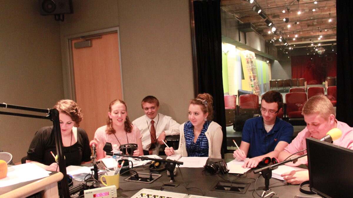 Six teens sitting behind microphones writing on pieces of paper on a radio studio