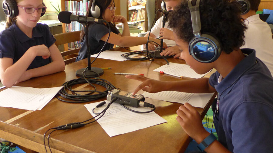 Five youth wearing headphones sitting behind microphones in a library