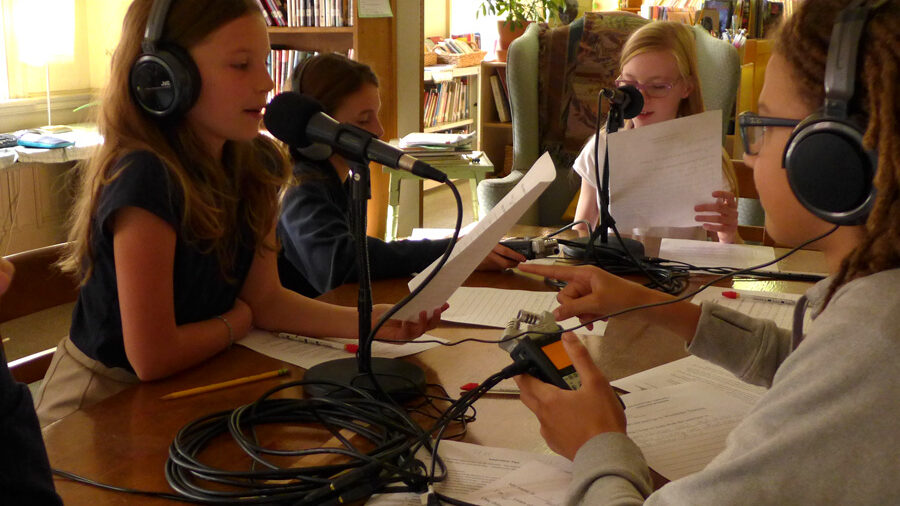 Four youth wearing headphones sitting behind microphones in a library