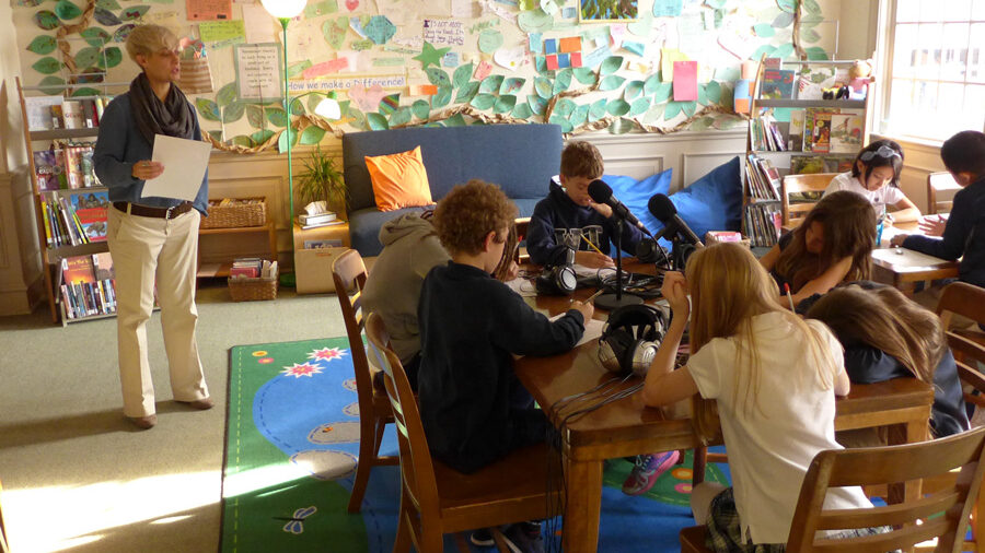 An adult looking at four youth sitting behind microphones in a library