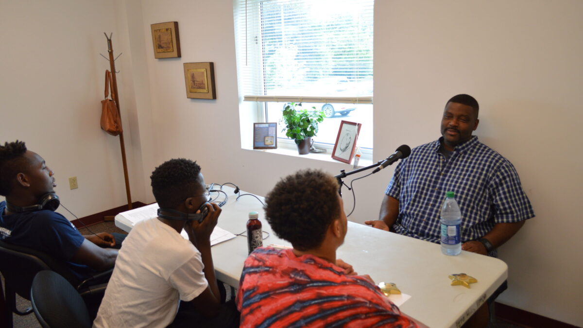 Three youth wearing headphones interviewing an adult speaking into a microphone.