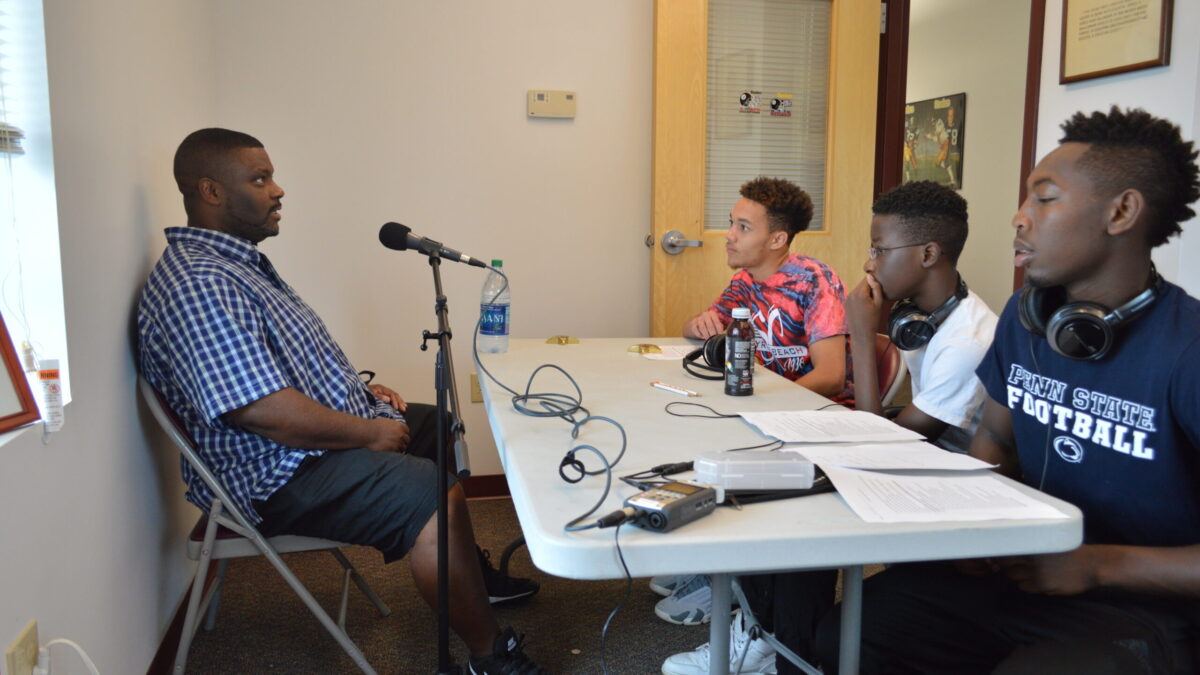 Three youth wearing headphones interviewing an adult speaking into a microphone.