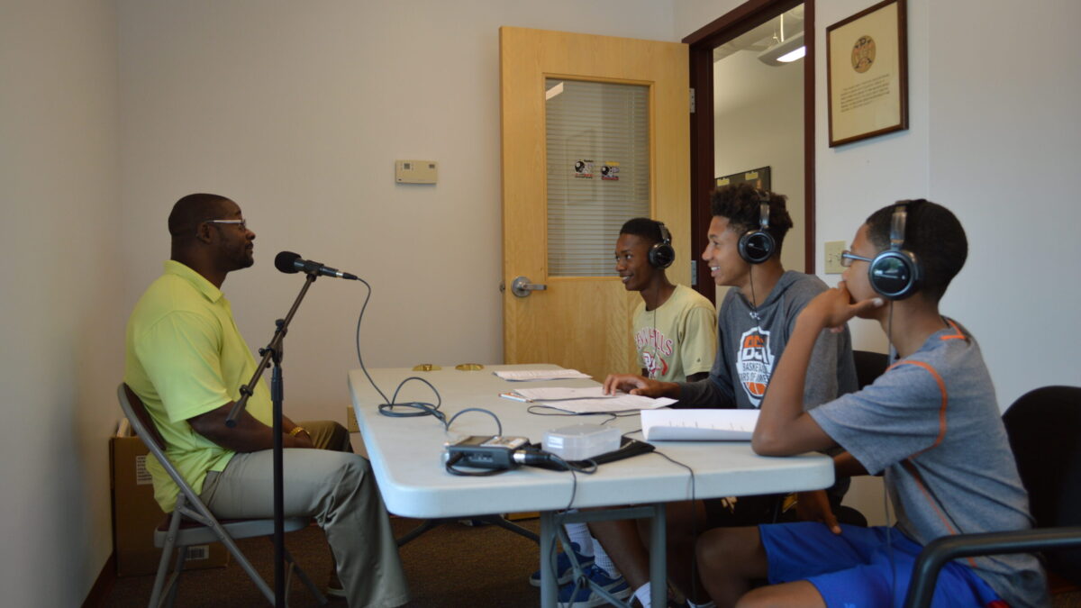 Three youth wearing headphones interviewing an adult speaking into a microphone.