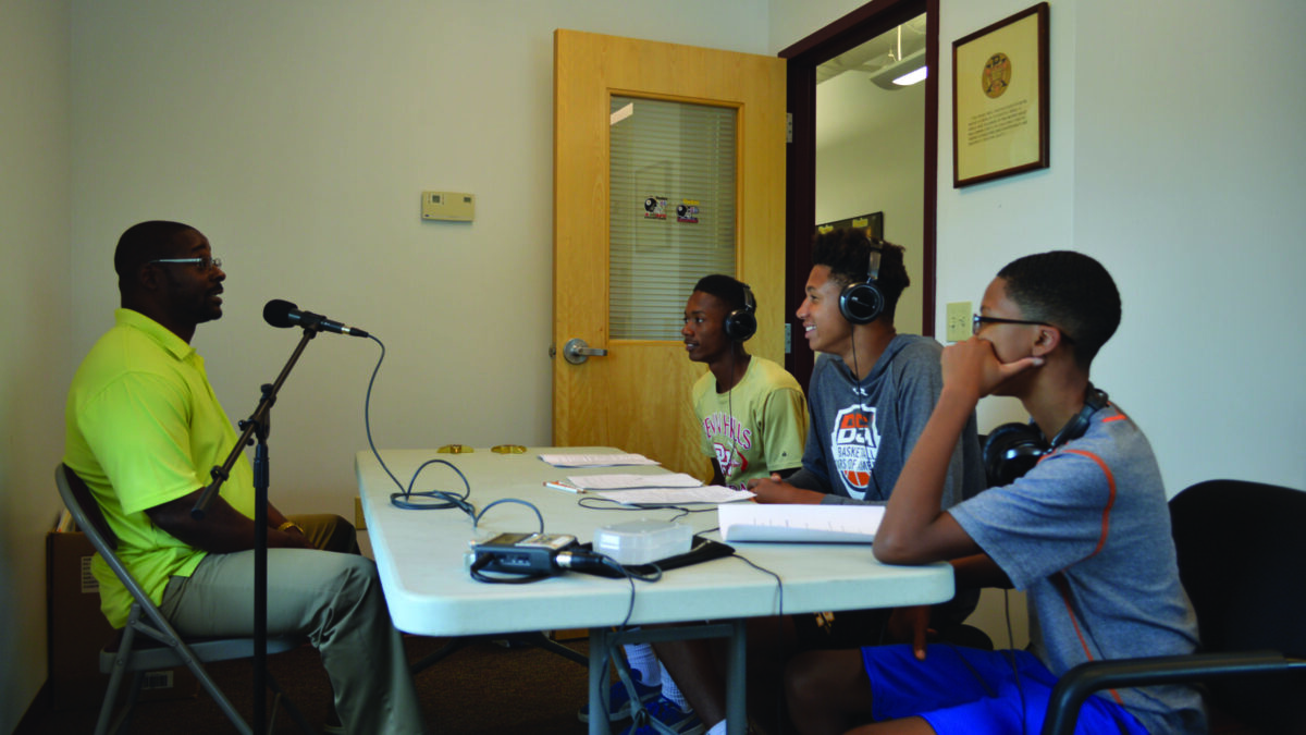 Three youth wearing headphones interviewing an adult speaking into a microphone.