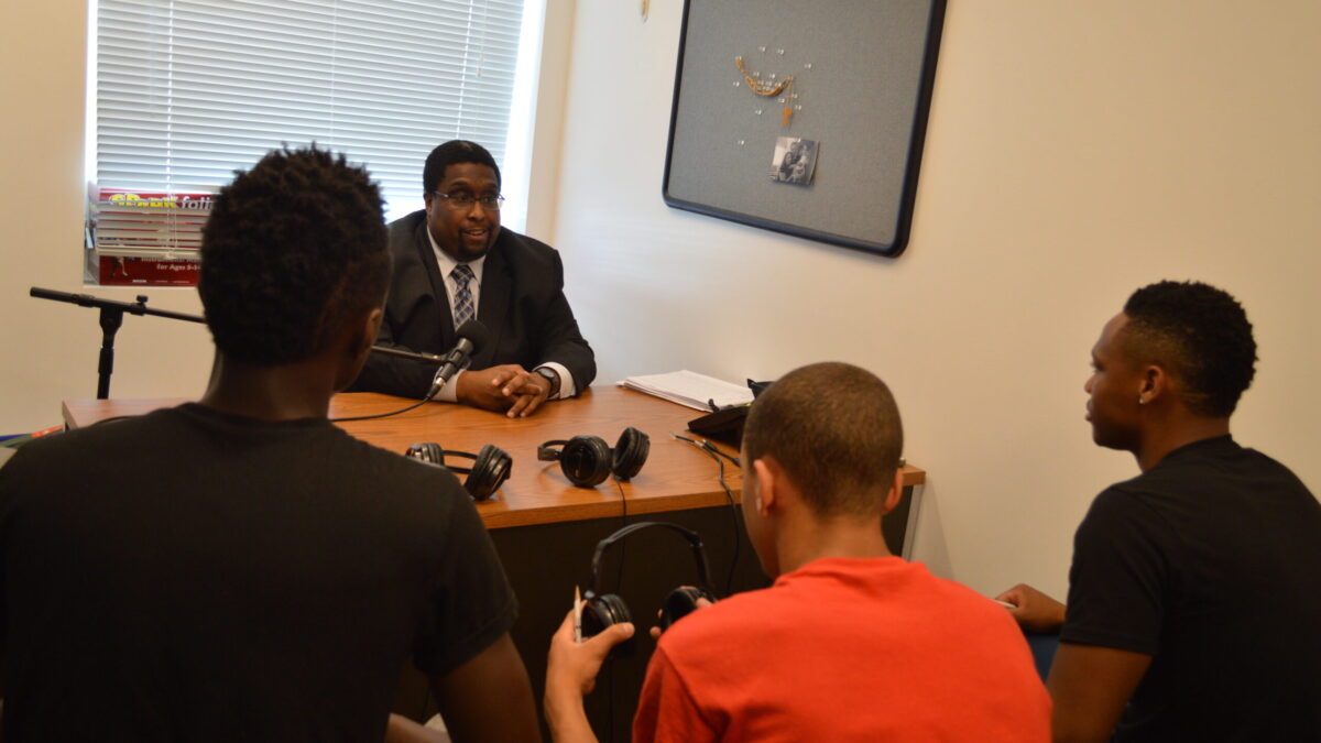 Three youth wearing headphones interviewing an adult speaking into a microphone.