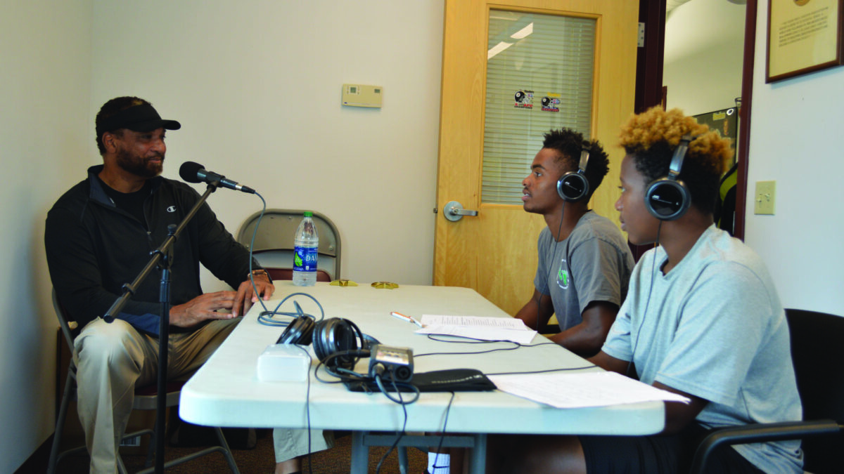 Two youth wearing headphones interviewing an adult speaking into a microphone.