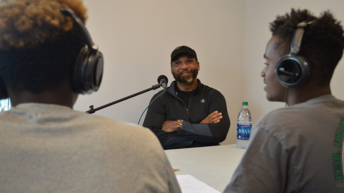 Two youth wearing headphones interviewing an adult speaking into a microphone.