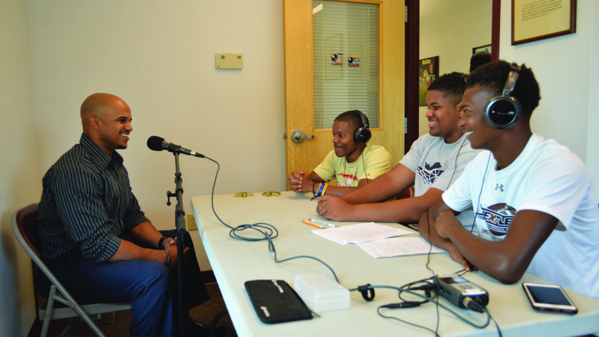 Three youth wearing headphones interviewing an adult speaking into a microphone.