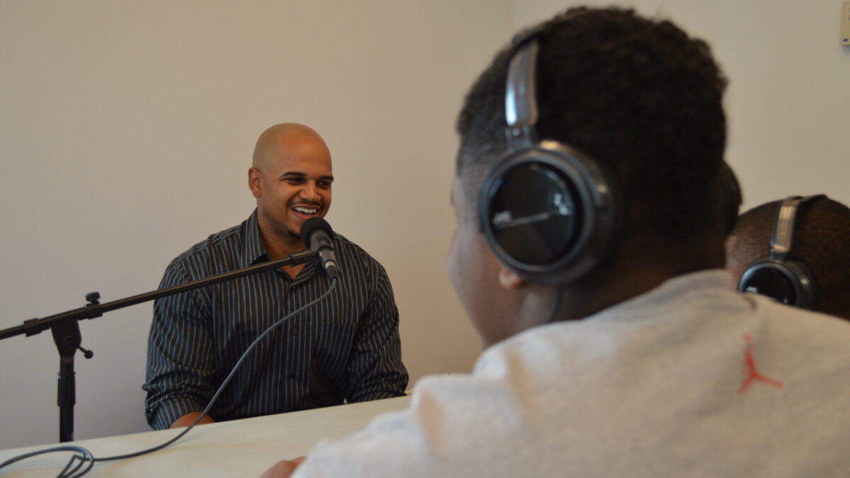 Two youth wearing headphones interviewing an adult speaking into a microphone.