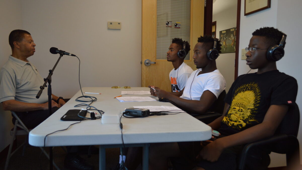 Three youth wearing headphones interviewing an adult speaking into a microphone.
