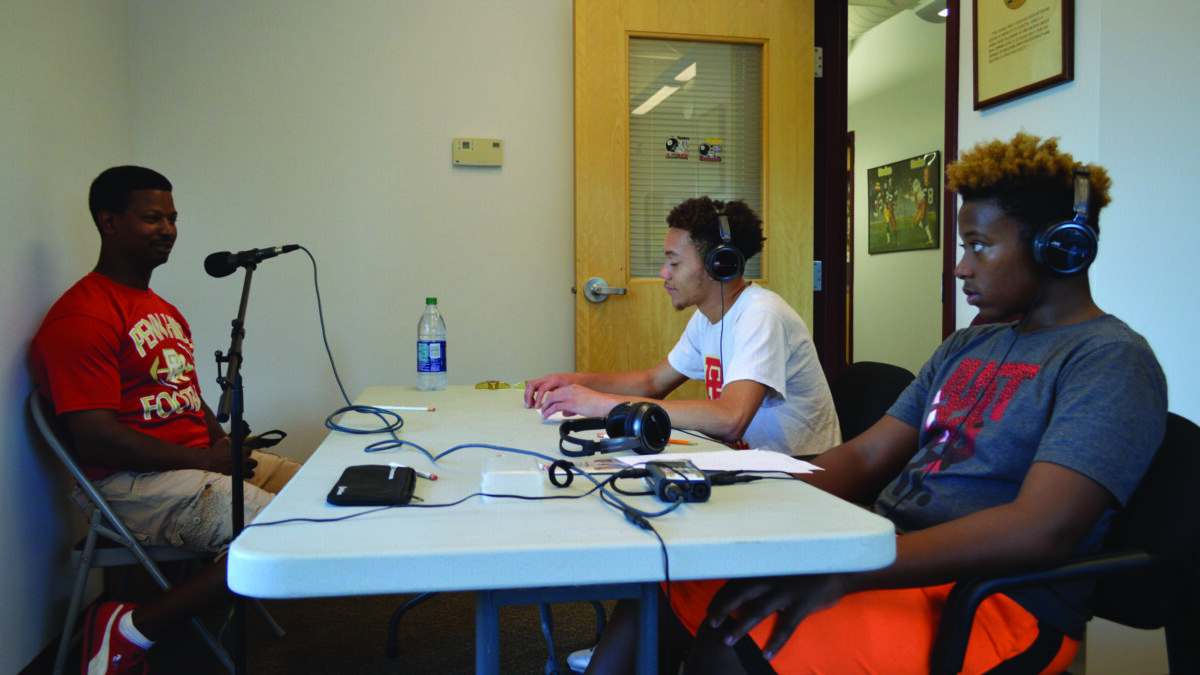 Two youth wearing headphones interviewing an adult speaking into a microphone.