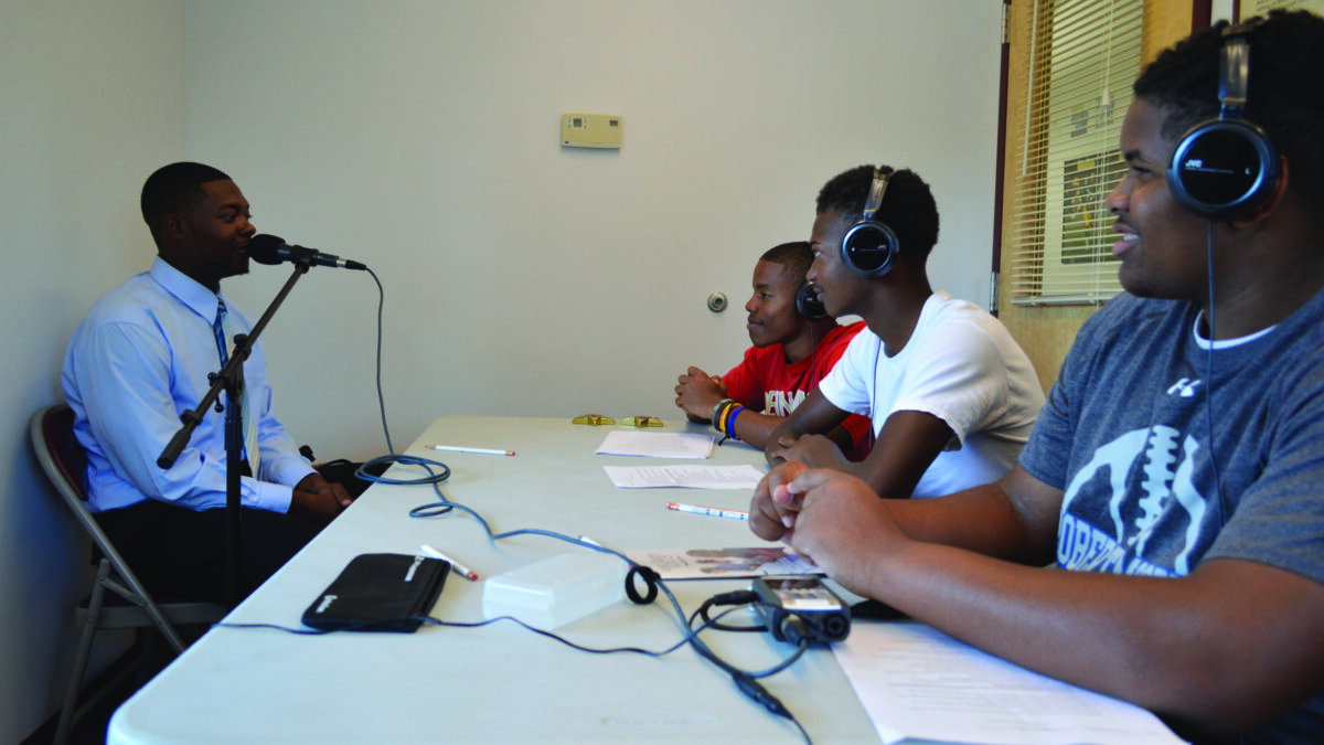 Three youth wearing headphones interviewing an adult speaking into a microphone.