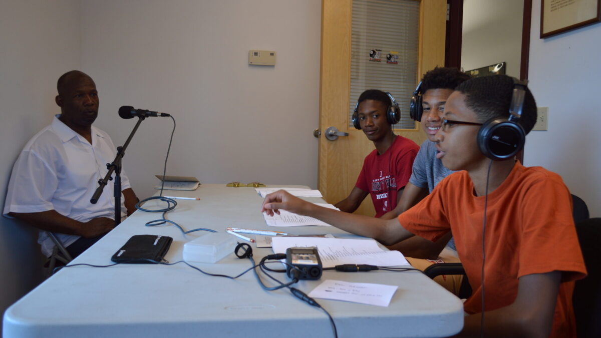 Three youth wearing headphones interviewing an adult speaking into a microphone.